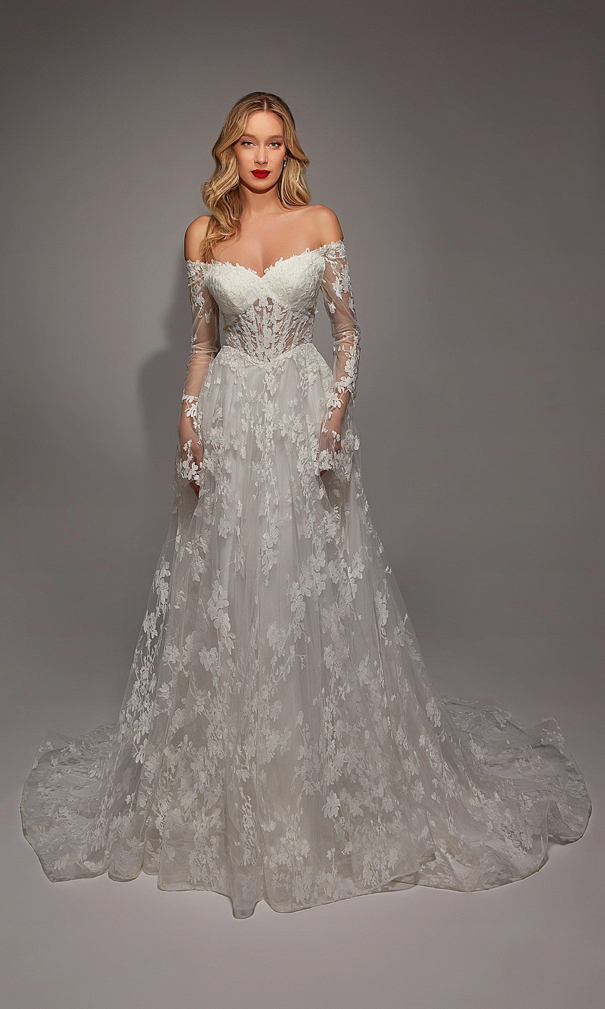 A woman stands in the Long White Dress: Alyce 7150, an off-the-shoulder, long-sleeved gown with lace and ruffle details and a flowing train, elegantly draped against a plain backdrop.